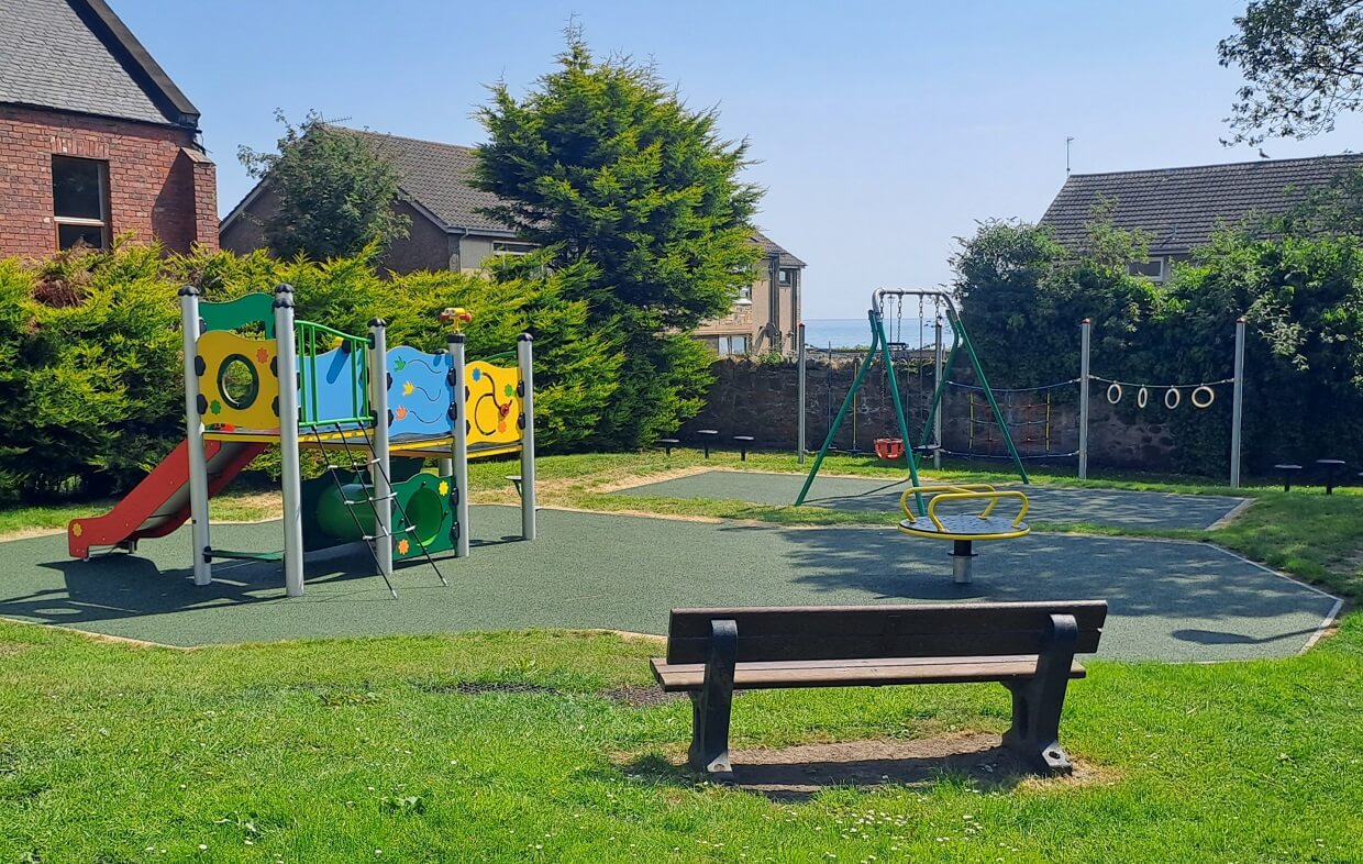 A playground with swings, slides, and climbing equipment. The playground is surrounded by green grass and houses in the background