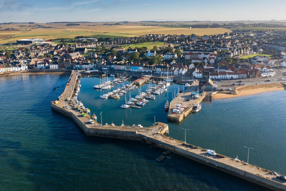 An aerial view of Anstruther's harbour area on a sunny day, taken from above the sea looking inland towards the town with the countryside stretching out into the distance.