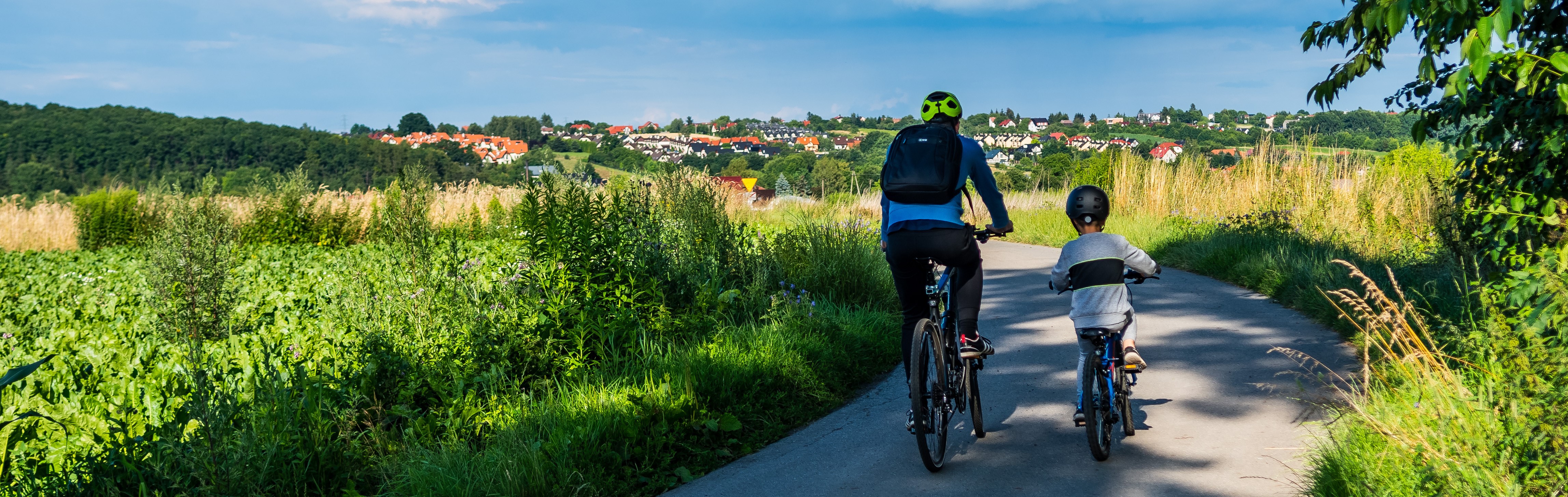  Two people are cycling on a paved path through a green, grassy area. A village is visible in the background under a blue sky. Two people are cycling on a paved path through a green, grassy area. A village is visible in the background under a blue sky.