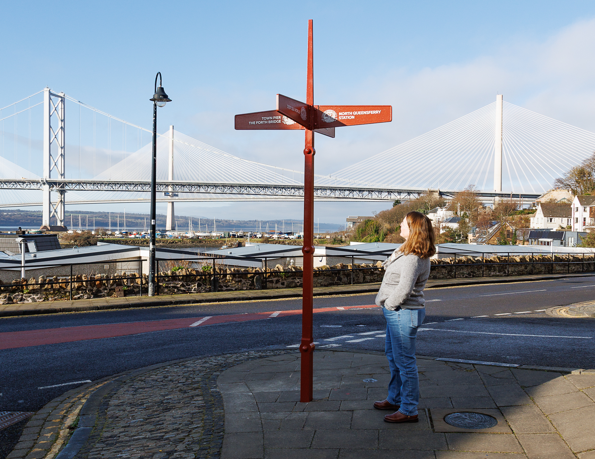 A woman looking up at the Forth Bridges Trail sign with a view of the Forth Bridges in the background.
