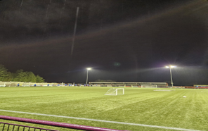  a brightly lit football field at night. Floodlights illuminate the green artificial turf. A white football goal is visible in the foreground.