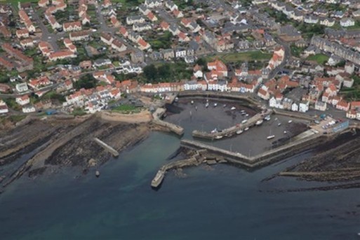 An aerial view of a St Monans featuring a harbor surrounded by breakwaters. The harbor contains several boats. The town is densely packed with houses and buildings, many with red-tiled roofs, extending inland from the waterfront. The coastline has rocky outcrops and the sea appears calm with varying shades of blue and green.