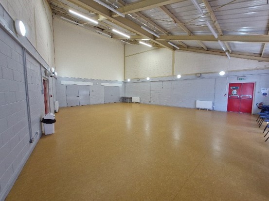 An empty indoor hall with a high ceiling and beige flooring. The walls are painted light gray, and there are several doors along the walls, including a red door on the right side. There are also some chairs stacked against the wall on the right side of the image. The hall is well-lit with multiple ceiling lights.