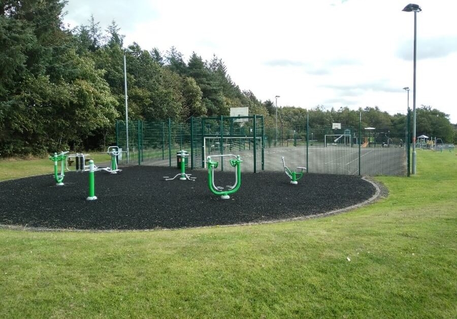 Fitness area at Gilvenbank Park with exercise equipment near sports courts and grassy fields.