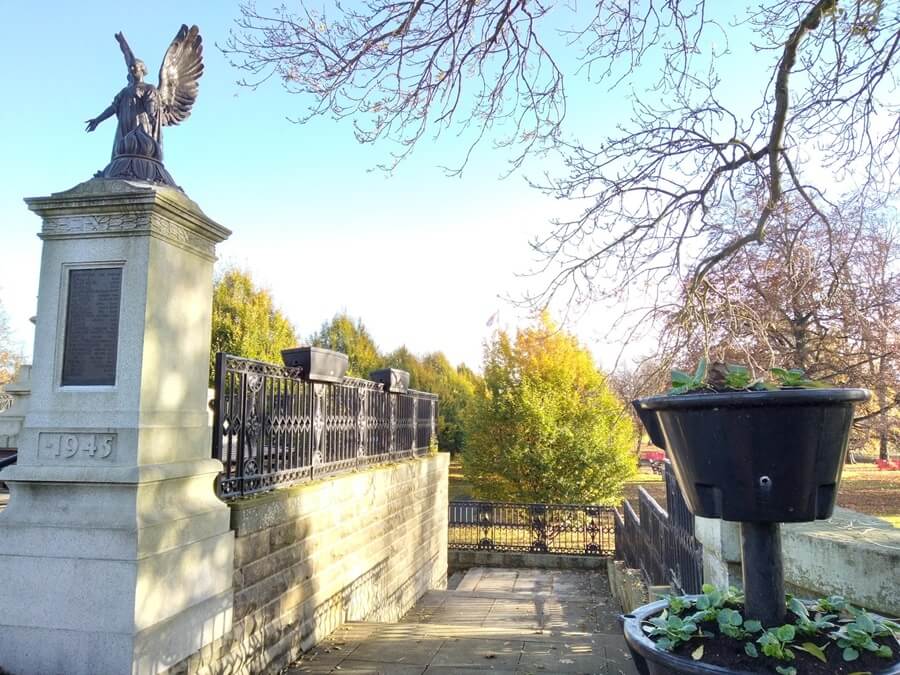 Statue of a bronze winged angel like sculpture atop a tall pedestal inscribed with '1945', surrounded by a black metal railing and steps, with autumn trees and a planter in the background