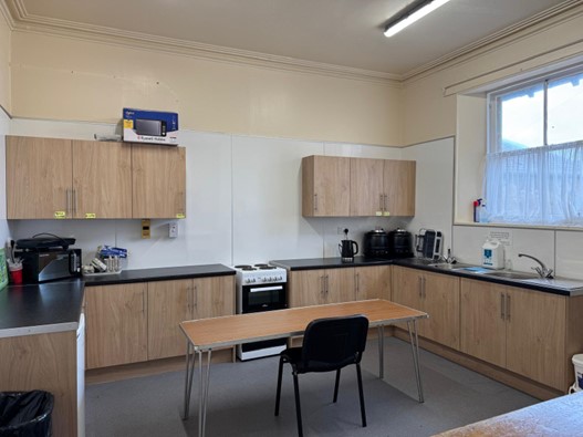 A spacious kitchen area with light wood cabinetry and black countertops, featuring an electric stove, a double sink under a window with a white curtain, and a free-standing wooden table with a single black chair in the centre.