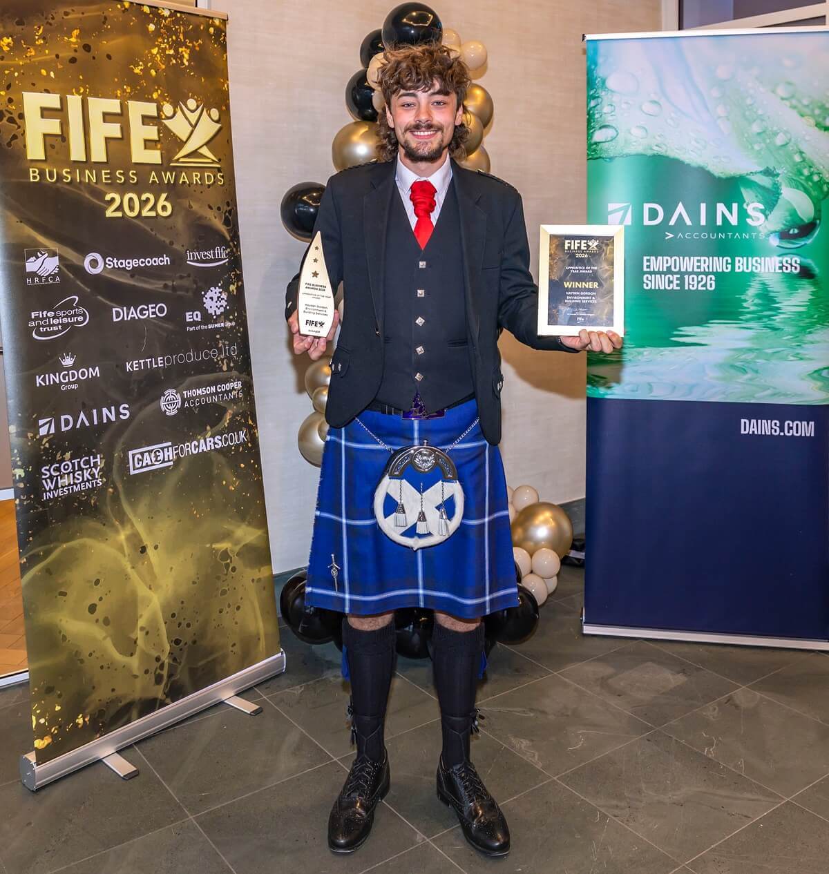 Person wearing a kilt and jacket holding two awards at the Fife Business Awards 2026, standing between sponsor banners and branded displays.