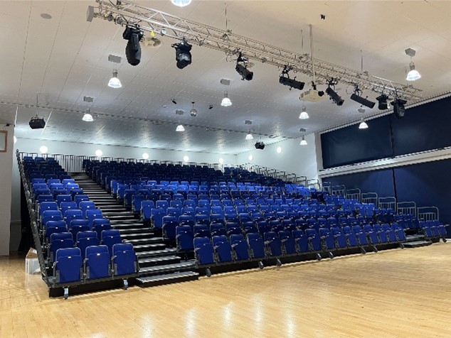 a tiered seating arrangement of blue chairs in a large hall with a wooden floor. Overhead, a metal truss supports various stage lights and equipment