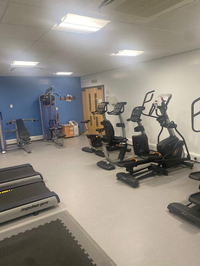 a fitness room with various exercise machines. There are treadmills in the foreground, followed by recumbent bikes and an elliptical trainer. In the background, there's a multi-station weight machine. The walls are painted in light blue and white, and the floor is covered with grey matting.
