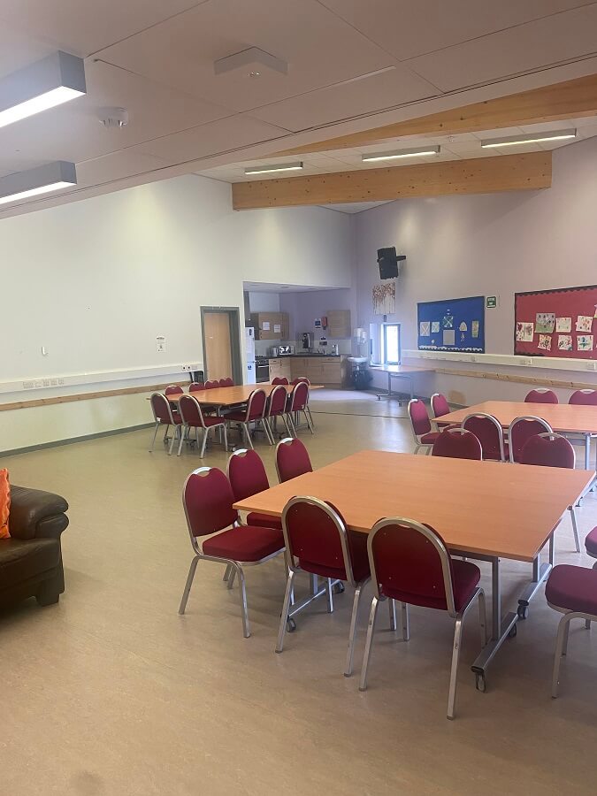 a room with tables and chairs. Some tables are arranged with red chairs around them, and there is a brown sofa in the corner. In the background, there's a serving area or kitchenette.