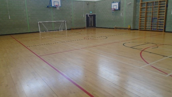 An indoor sports hall with court lines. Basketball hoops and a football net are visible. 