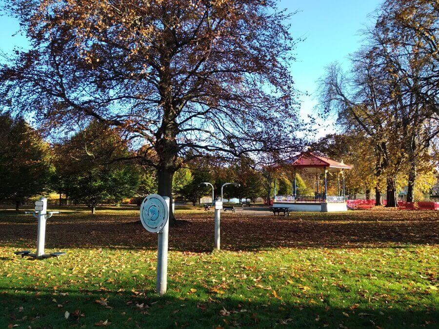 Haugh Park with outdoor exercise equipment, a large tree, and a red-roofed bandstand among fallen leaves