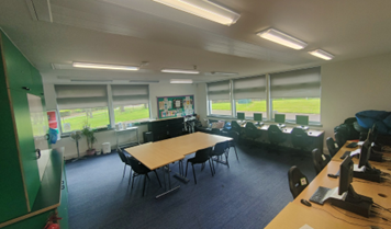 A wide shot of a modern IT training room with blue carpet and large windows. Several computer workstations line the right wall, and a long rectangular table with chairs is positioned in the center.