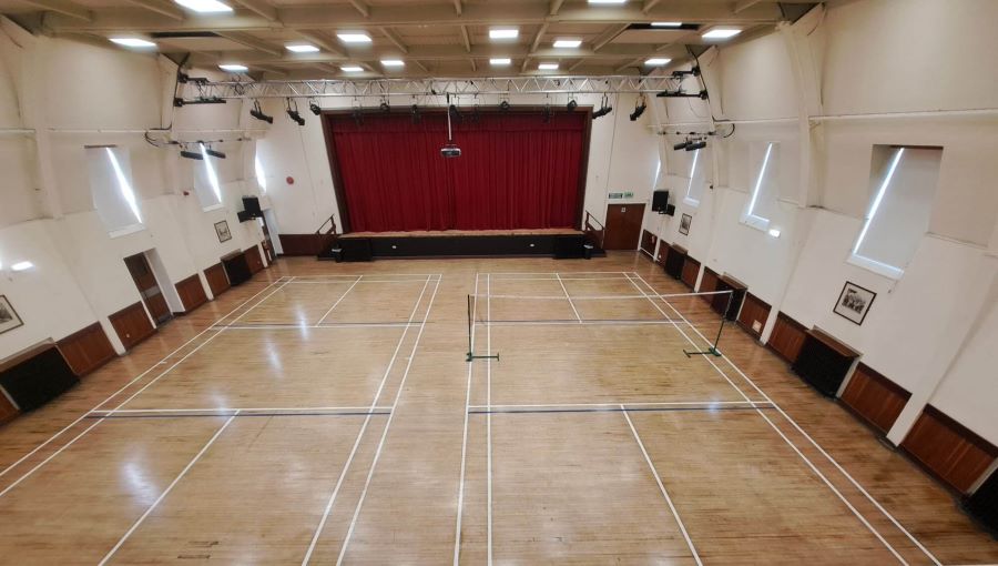 Large indoor hall with a polished wooden floor marked for badminton courts, a badminton net in the centre, and a stage with red curtains at the far end