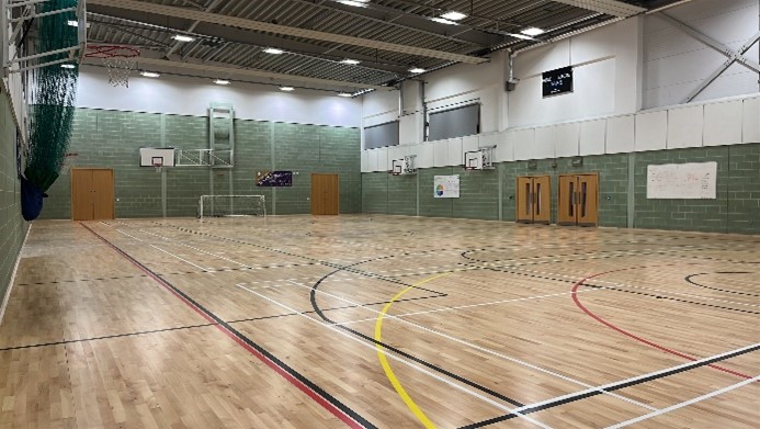 A brightly lit indoor gymnasium with a polished wooden floor marked with colourful lines for various sports. Basketball hoops are visible at both ends of the hall, and large double doors are located along the side walls. The walls are a combination of light green and white brick.