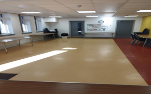 a spacious room with a light brown linoleum floor. There are several tables with chairs arranged in different areas of the room. Natural light enters through windows on the left wall. There are doorways on the back wall, and a whiteboard with papers is visible on the right side of the back wall. Part of a red floor is visible in the right corner.