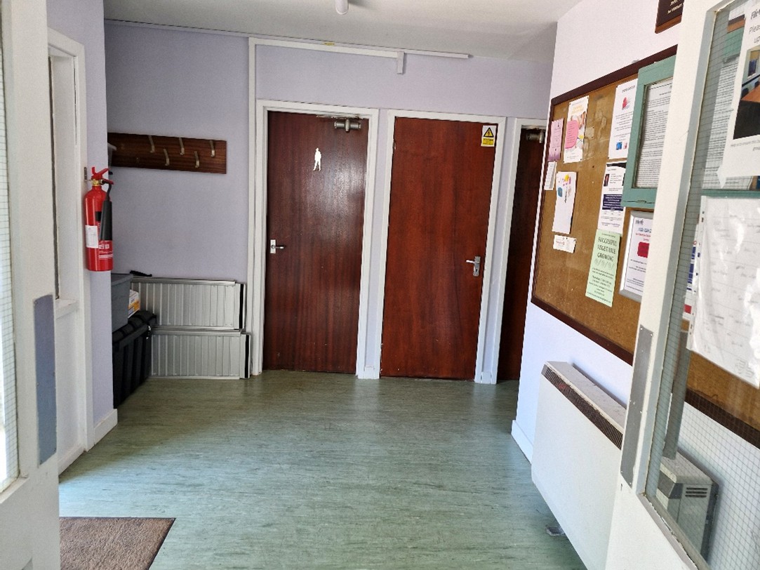 A hallway with two wooden doors, a fire extinguisher mounted on the wall, and a bulletin board with various notices and papers pinned to it. There is also a coat rack with hooks on the left wall and some storage containers below it. The floor is covered in light green linoleum, and there is a radiator on the right side of the hallway.