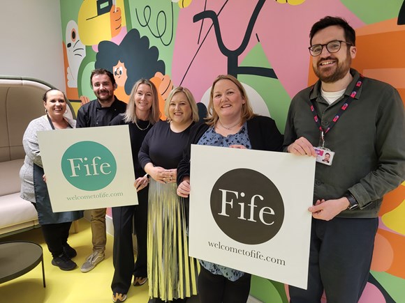 A group of six people hold up two 'Welcome to Fife' branded boards, everyone is smiling at the camera. They are staff from the Welcome to Fife team and Adam Smith Theatre in Kirkcaldy.