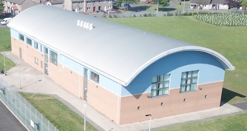 An aerial view shows a modern, curved building with a silver roof, light blue and brick walls, and large windows. The building is surrounded by green grass and a few trees.