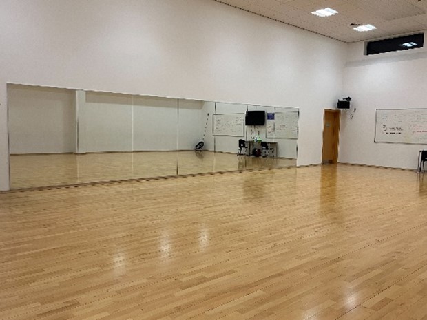  a dance studio with a polished wooden floor and white walls. A large mirror covers a significant portion of one wall, reflecting the studio space. There are also whiteboards and some equipment visible in the background