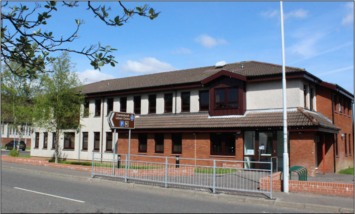 the Maxwell Centre in Cowdenbeath. It is a two-story building with a brown tiled roof and a mix of brick and light-colored walls. A sign points towards the "Cowdenbeath Leisure Centre" and indicates parking. A street and a metal railing are in the foreground, and trees and a blue sky with some clouds are visible in the background
