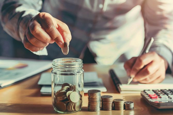 Image shows a desk with a jar filled with coins, some stacks of coins, a calculator, some papers and a person's hand putting money into the jar