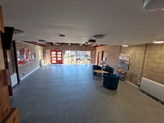 A spacious, well-lit room with a high ceiling and large windows at the far end, allowing natural light to enter. The room has a polished floor and is furnished with three blue armchairs and a small round table in the center-right area. On the left wall, there are several bulletin boards with various notices and posters. The right wall has shelves filled with books or magazines. There are also radiators along both walls for heating.