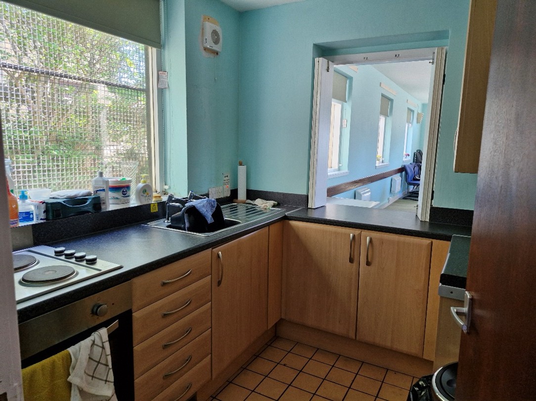A small kitchen with light blue walls and wooden cabinets. There is a window on the left side with various items on the windowsill, including cleaning supplies and a dish rack. The countertop has a sink with dishes in it, an electric stove, and other kitchen essentials. There is an open serving hatch that looks into another room with several windows and chairs visible. The floor is tiled in a brown color.