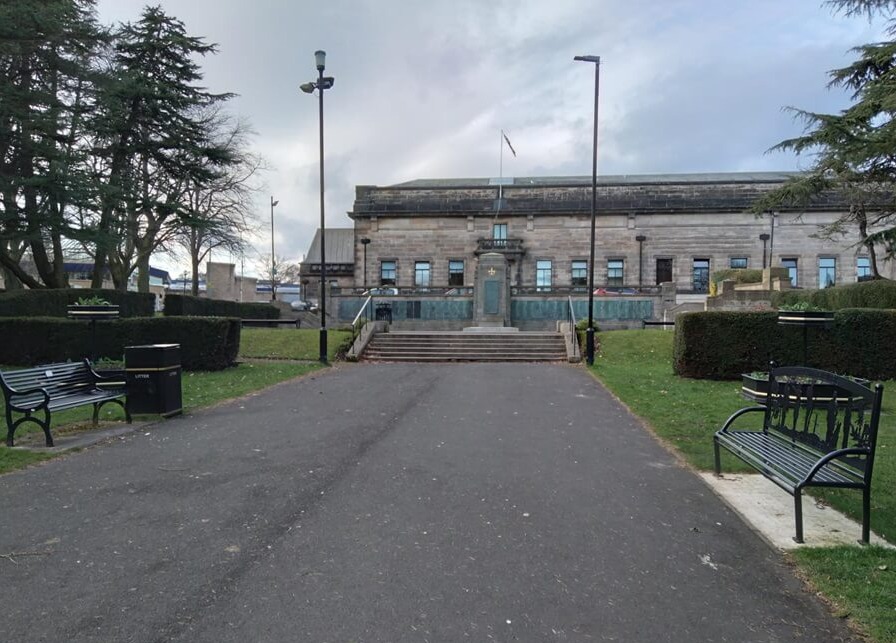 Pathway with benches and trees leading to a large stone building with stairs and a flagpole on the roof