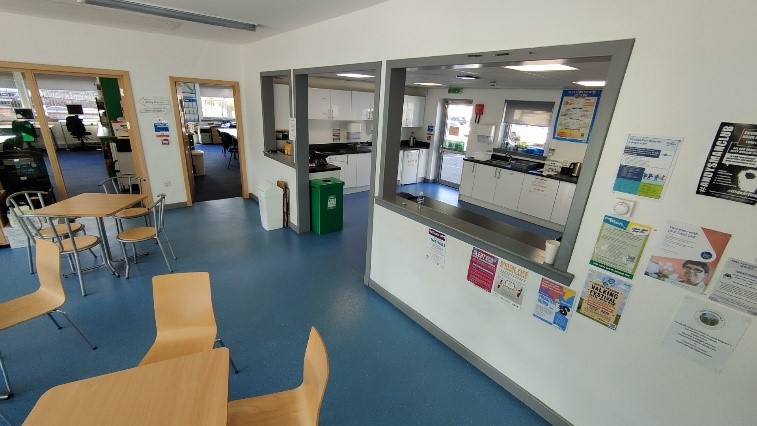 A large, bright room with a blue floor, functioning as both a dining area and a view into a kitchen. Several light wood tables with matching chairs are in the foreground, and a serving counter with an open pass-through looks into a white kitchen area beyond. Various notices and posters are taped to the wall on the right.