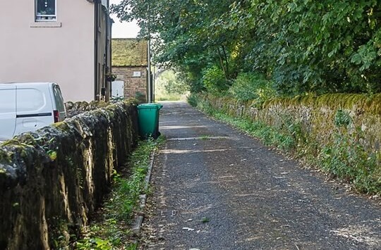 Narrow Woolmill Road in residential area between trees and stone wall - £30,000 approved for Woolmill Road improvements