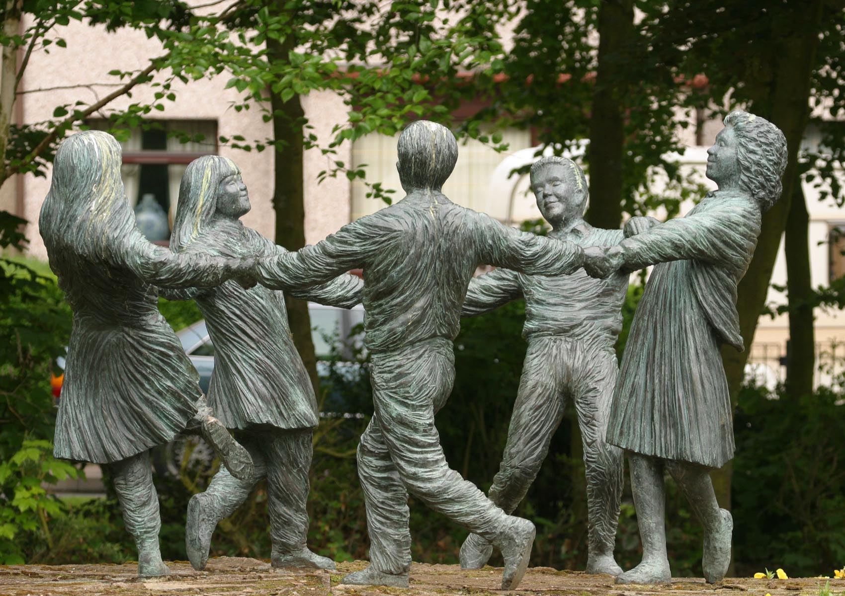 A bronze sculpture of six children holding hands and dancing in a circle. The sculpture is outdoors with trees in the background