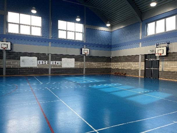 a gymnasium with a blue floor and blue brick walls. There are basketball hoops on the walls, and whiteboards are visible on the left side