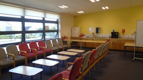  An indoor community room. There are rows of chairs and tables. A small kitchen area is visible