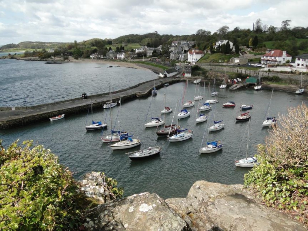 A coastal scene featuring a Aberdour harbour with numerous sailboats anchored in the water. The harbour is bordered by a stone pier that extends into the water, and there are several buildings, including houses, situated on the hillside overlooking the harbour. The landscape in the background includes rolling hills and greenery.