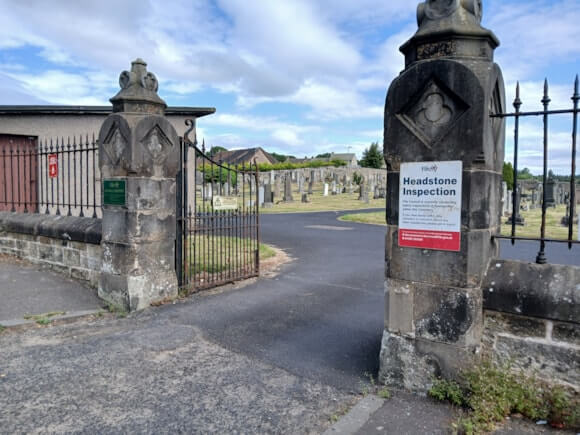 Entrance to Bowhill Cemetery, showing open wrought-iron gates between two stone gateposts. A sign on the right-hand pillar reads “Headstone Inspection,” while rows of gravestones and a paved pathway are visible inside the cemetery under a partly cloudy blue sky.