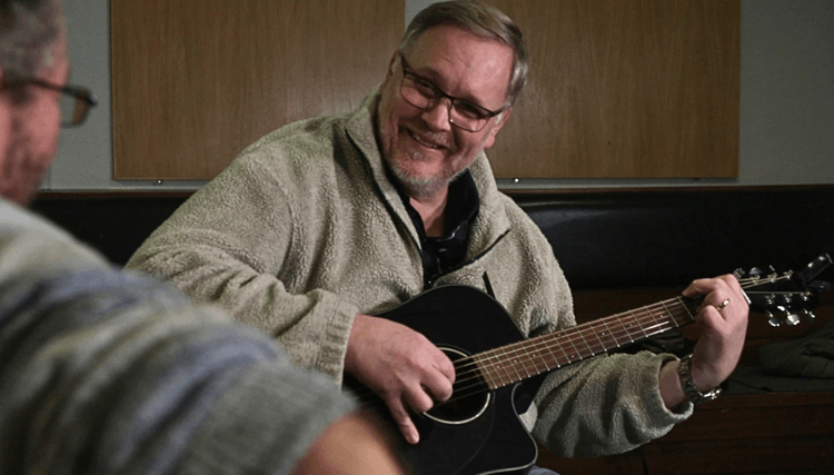 A person sits indoors holding and playing a black acoustic guitar during a lesson or group music session. Another participant is visible out of focus in the foreground, also holding a guitar. The setting has wooden wall panels and soft lighting, creating an informal learning environment.