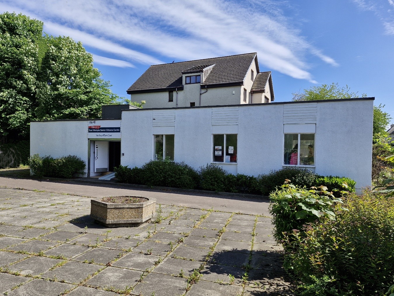 A building with a white exterior and three visible windows. There is a sign above the entrance that reads 'Welcome to East Wemyss Senior Citizens Centre' The entrance door is located on the left side of the building, and there are bushes in front of the building. In front of the building, there is a paved area with a circular planter in the center. There are trees and another house visible in the background.