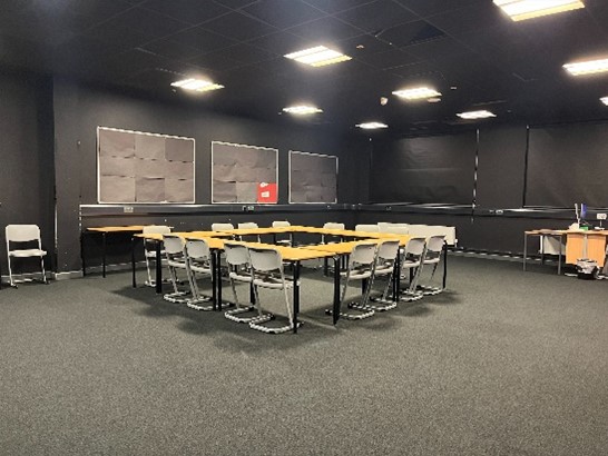 a classroom or meeting room with dark walls and a dark carpet. A square arrangement of tables and chairs is in the center of the room. There are also individual tables and chairs along the walls, and some papers are pinned to the wall at the back. The room is lit by several ceiling lights