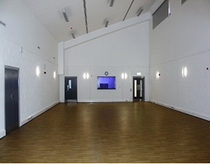 a large, brightly lit hall with a polished wooden floor. The walls are white, and there are several doors and a small window visible.