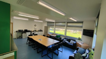  A long, rectangular multi-purpose room with a blue floor. A large conference table with chairs occupies the center, while a black sofa sits near windows on the right. A counter with various equipment lines the back wall, and bright fluorescent lights illuminate the ceiling.