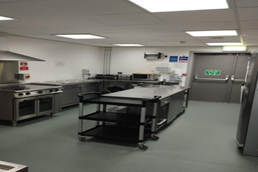 a commercial kitchen with stainless steel appliances, including ovens, a stovetop, and work surfaces. A mobile cart with shelves is in the center of the room. The floor is grey, and there is an exit door on the right side.