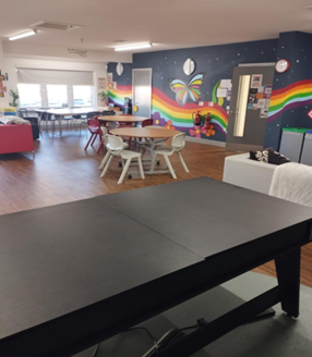a room with a black table in the foreground. In the background, there are round tables with white and red chairs, and a colorful mural on the wall.