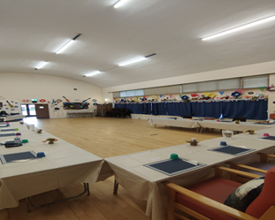 A wide shot of a community hall with light wood flooring. Several rectangular tables, covered with white tablecloths and set with place settings, are arranged in a U-shape. Blue curtains hang on the far wall, decorated with colorful paper shapes.