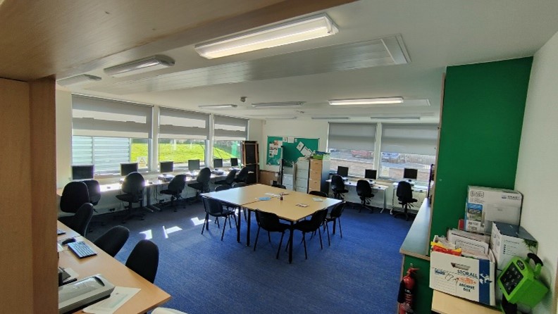 A brightly lit meeting room with a dark blue carpet. Several computer workstations are arranged along the far wall beneath large windows. In the center of the room, a rectangular table with chairs is set up for meetings.