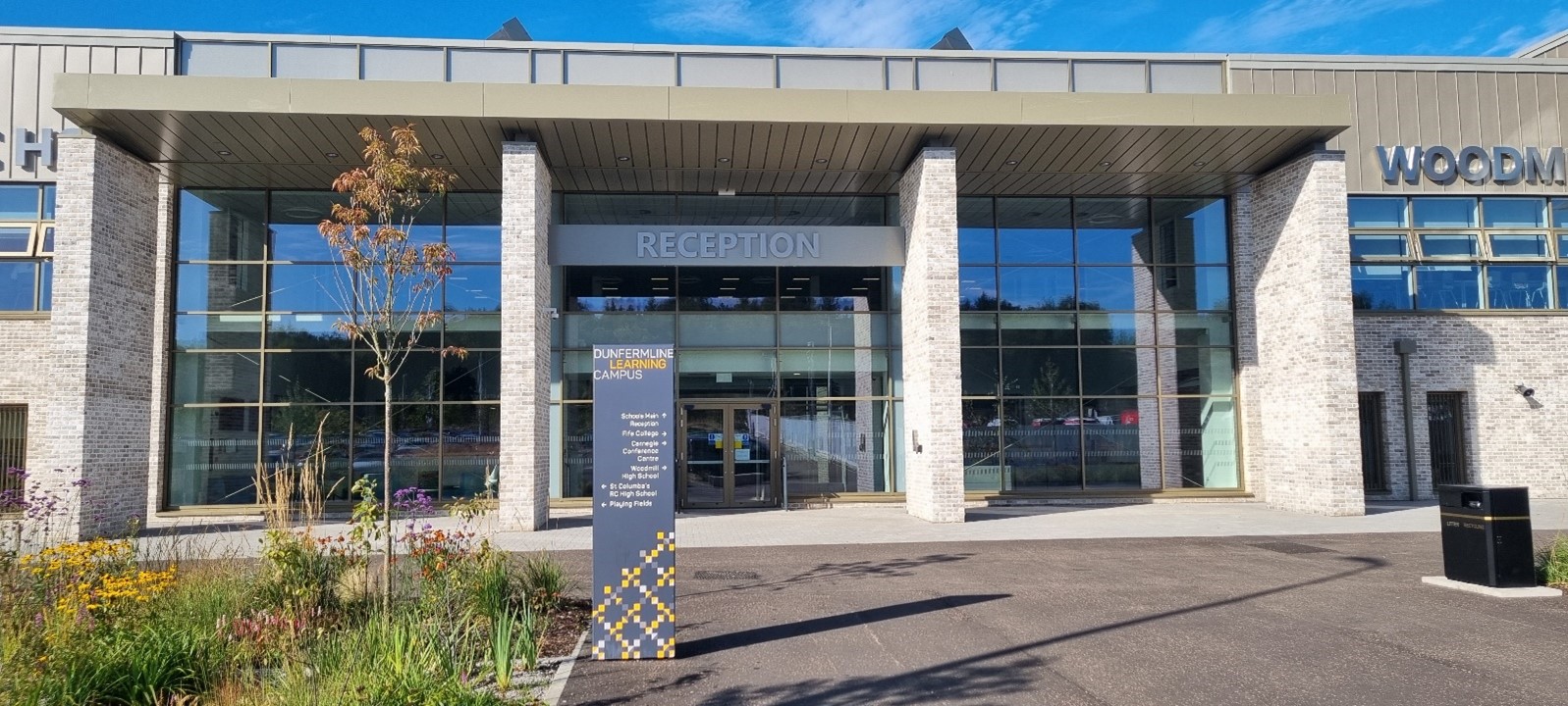  A grand modern building entrance with a wide glass facade and large stone pillars. The word "RECEPTION" is visible above the main doors. A tall, dark sign with yellow text and a patterned base stands in front of the entrance, providing directions.