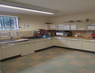  A functional kitchen with light-colored cabinets and countertops forming an L-shape. A sink is visible on the left, and a microwave sits on the counter to the right. The floor has a checkered pattern of light and dark tiles.