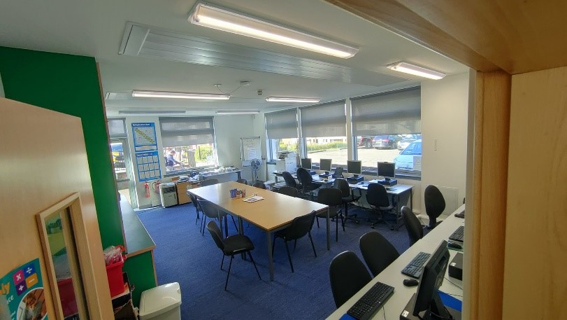  A meeting room with blue carpet and large windows. Computer workstations are set up along the right wall. A long conference table with chairs is in the center of the room. Various posters and charts are on the walls.
