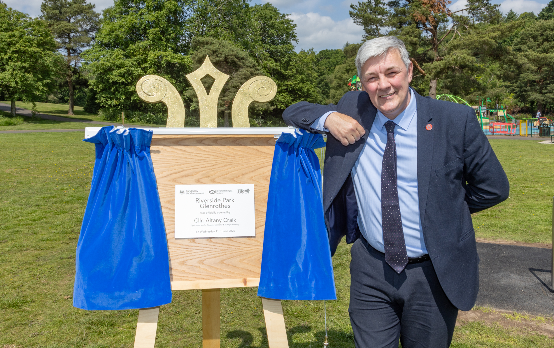 Cllr Altany Craik standing at the opening ceremony of Riverside Park, trees, and a play park are in the background.