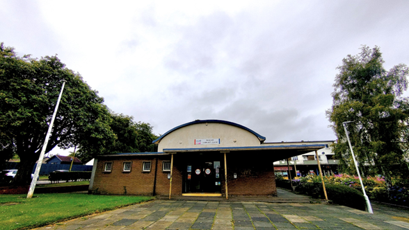A low-angle exterior shot shows a squat, brown brick building with a curved, light-colored roof. A flat awning extends over the entrance, supported by yellow poles. Green trees flank the building on either side, and the sky is overcast